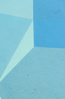 An abstract aerial view of a geometric pattern composed of large, flat, interconnected surfaces. The colors vary in shades of blue and teal, creating a visually interesting mosaic with lines forming a tessellated pattern across the surface. Tiny, cone-shaped objects project upwards at regular intervals, adding texture and depth to the scene.