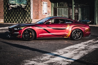 A sleek, customized sports car in vibrant red with black and yellow accents is parked in an urban setting. The car features sporty enhancements and is positioned in front of a brick building with artistic murals and posters, adding a creative backdrop.