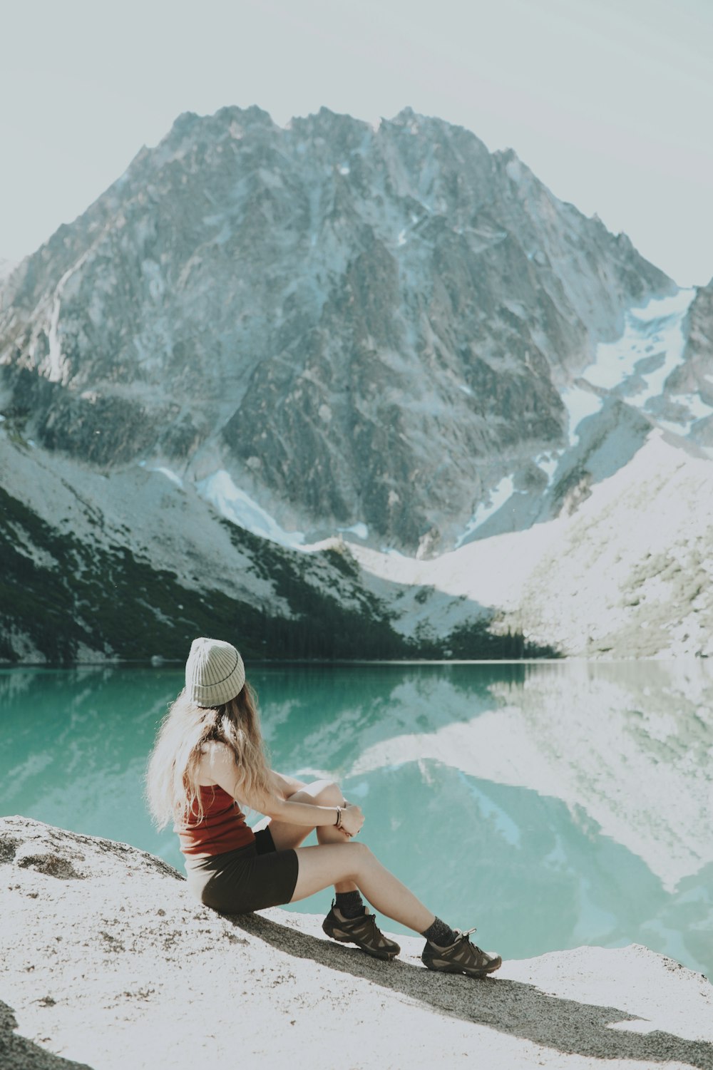 Mujer con camiseta sin mangas negra y gorra de punto blanca sentada en una roca cerca del lago durante el día