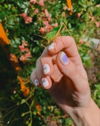 A close-up of a hand with manicured nails featuring minimalist designs, gently holding a green grasshopper. The background contains blurred greenery and pink flowers.