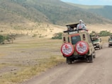 Two safari vehicles drive along a dirt road in a vast, open landscape. The area is surrounded by rolling hills with sparse vegetation. Several people stand in the open sunroofs of the vehicles, enjoying the view, with one person taking a photograph.