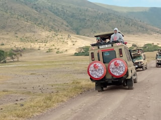 Two safari vehicles drive along a dirt road in a vast, open landscape. The area is surrounded by rolling hills with sparse vegetation. Several people stand in the open sunroofs of the vehicles, enjoying the view, with one person taking a photograph.