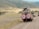 Two safari vehicles drive along a dirt road in a vast, open landscape. The area is surrounded by rolling hills with sparse vegetation. Several people stand in the open sunroofs of the vehicles, enjoying the view, with one person taking a photograph.