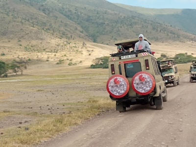 Two safari vehicles drive along a dirt road in a vast, open landscape. The area is surrounded by rolling hills with sparse vegetation. Several people stand in the open sunroofs of the vehicles, enjoying the view, with one person taking a photograph.