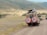 Two safari vehicles drive along a dirt road in a vast, open landscape. The area is surrounded by rolling hills with sparse vegetation. Several people stand in the open sunroofs of the vehicles, enjoying the view, with one person taking a photograph.