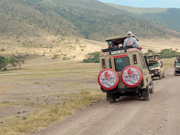 Two safari vehicles drive along a dirt road in a vast, open landscape. The area is surrounded by rolling hills with sparse vegetation. Several people stand in the open sunroofs of the vehicles, enjoying the view, with one person taking a photograph.