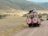 Two safari vehicles drive along a dirt road in a vast, open landscape. The area is surrounded by rolling hills with sparse vegetation. Several people stand in the open sunroofs of the vehicles, enjoying the view, with one person taking a photograph.