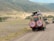 Two safari vehicles drive along a dirt road in a vast, open landscape. The area is surrounded by rolling hills with sparse vegetation. Several people stand in the open sunroofs of the vehicles, enjoying the view, with one person taking a photograph.