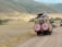 Two safari vehicles drive along a dirt road in a vast, open landscape. The area is surrounded by rolling hills with sparse vegetation. Several people stand in the open sunroofs of the vehicles, enjoying the view, with one person taking a photograph.