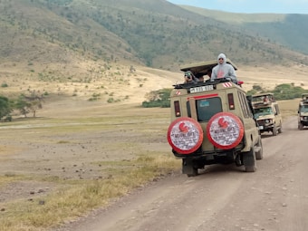 Two safari vehicles drive along a dirt road in a vast, open landscape. The area is surrounded by rolling hills with sparse vegetation. Several people stand in the open sunroofs of the vehicles, enjoying the view, with one person taking a photograph.