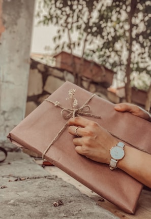 A pair of hands delicately hold a wrapped gift tied with twine and adorned with small flowers. The person wears a wristwatch and a ring, suggesting a sense of anticipation and care. The background features foliage and a rustic outdoor setting, adding to the serene and thoughtful atmosphere.