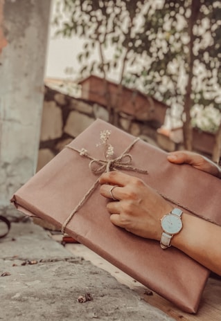 A pair of hands delicately hold a wrapped gift tied with twine and adorned with small flowers. The person wears a wristwatch and a ring, suggesting a sense of anticipation and care. The background features foliage and a rustic outdoor setting, adding to the serene and thoughtful atmosphere.