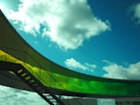 A modern architectural structure with a curved, semi-transparent yellow and green glass railing against a clear blue sky with scattered clouds. An industrial metal staircase with handrails leads up to the structure.