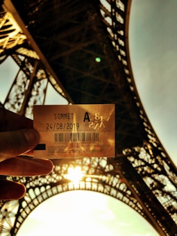 A hand holds a ticket with the date '24/08/2019' against the backdrop of the Eiffel Tower. The sun is shining brightly, creating a silhouette effect and illuminating the intricacies of the tower's iron lattice structure.