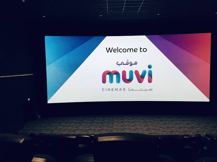 A movie theater interior with rows of empty seats facing a large screen displaying a greeting message for Muvi Cinemas. The screen is brightly lit with a combination of blue, red, and white colors.