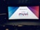 A movie theater interior with rows of empty seats facing a large screen displaying a greeting message for Muvi Cinemas. The screen is brightly lit with a combination of blue, red, and white colors.