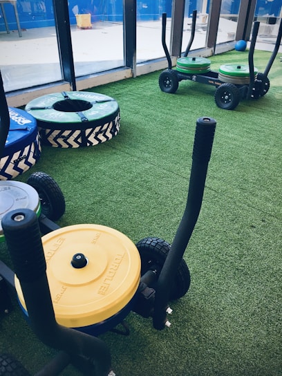 A gym setting with equipment used for weight training resting on artificial grass. There are weighted sleds with handles, featuring colorful plates stacked on them, including yellow and green ones. Additional training apparatuses like large round cushions or bumpers with blue and green covers are visible, designed for strength workouts. The room is bordered by large windows allowing natural light to enter.