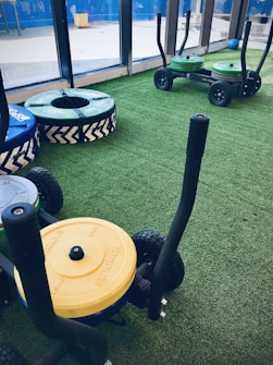 A gym setting with equipment used for weight training resting on artificial grass. There are weighted sleds with handles, featuring colorful plates stacked on them, including yellow and green ones. Additional training apparatuses like large round cushions or bumpers with blue and green covers are visible, designed for strength workouts. The room is bordered by large windows allowing natural light to enter.