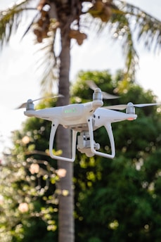 A white drone hovers in the air with a blurred background of a tall palm tree and lush green foliage.
