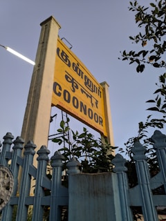 A yellow sign with text in multiple languages is prominently displayed, surrounded by lush green foliage. The sign is mounted on a tall post beside a light blue fence. The sky in the background is clear and subtly tinted, suggesting late afternoon or early evening.