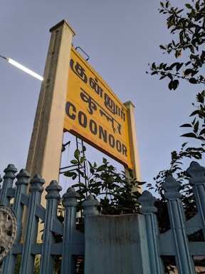 A yellow sign with text in multiple languages is prominently displayed, surrounded by lush green foliage. The sign is mounted on a tall post beside a light blue fence. The sky in the background is clear and subtly tinted, suggesting late afternoon or early evening.