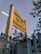 A yellow sign with text in multiple languages is prominently displayed, surrounded by lush green foliage. The sign is mounted on a tall post beside a light blue fence. The sky in the background is clear and subtly tinted, suggesting late afternoon or early evening.
