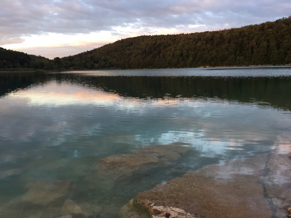 lake near green trees and mountain under cloudy sky during daytime