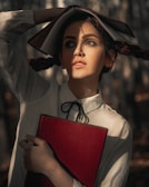 A woman with expressive makeup holds a red book against her chest. Her hair is styled in pigtails, and she has another book open on her head as if using it for shade. The sunlight creates dramatic shadows on her face, giving the scene an artistic and contemplative feel. She wears a white blouse with a black ribbon tie.
