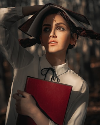 A woman with expressive makeup holds a red book against her chest. Her hair is styled in pigtails, and she has another book open on her head as if using it for shade. The sunlight creates dramatic shadows on her face, giving the scene an artistic and contemplative feel. She wears a white blouse with a black ribbon tie.
