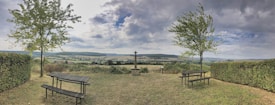 A serene landscape featuring two picnic tables positioned on a grassy area, bordered by neatly trimmed hedges. Two tall trees stand on either side, framing the scene. In the distance, rolling hills stretch under a sky filled with a mix of clouds and patches of blue. A cross mounted on a stone pedestal adds a focal point to the tranquil rural vista.