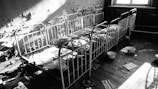 A series of rusty metal bed frames are lined against a decayed wall with peeling paint. The room appears abandoned, with scattered sheets of paper and enamel basins on the floor. Light from a window casts shadows across the beds, enhancing the feeling of neglect and abandonment.