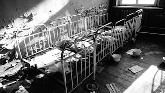 A series of rusty metal bed frames are lined against a decayed wall with peeling paint. The room appears abandoned, with scattered sheets of paper and enamel basins on the floor. Light from a window casts shadows across the beds, enhancing the feeling of neglect and abandonment.