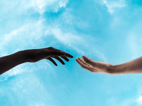 Two hands reaching towards each other against a bright blue sky with scattered clouds. The composition captures a sense of connection and reaching out.