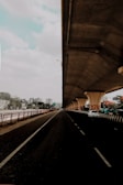 A cityscape featuring a long, empty road beneath a large, elevated highway or bridge. The road stretches into the distance, flanked by railings and greenery. On the right side, there is a parked white car under the bridge's supportive pillars, which are decorated with a geometric pattern. High-rise buildings are visible in the background under a partly cloudy sky.
