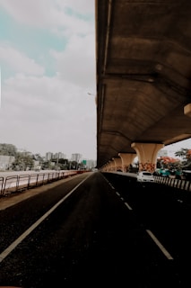 A cityscape featuring a long, empty road beneath a large, elevated highway or bridge. The road stretches into the distance, flanked by railings and greenery. On the right side, there is a parked white car under the bridge's supportive pillars, which are decorated with a geometric pattern. High-rise buildings are visible in the background under a partly cloudy sky.