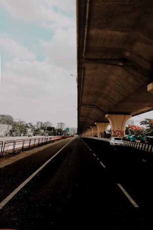 A cityscape featuring a long, empty road beneath a large, elevated highway or bridge. The road stretches into the distance, flanked by railings and greenery. On the right side, there is a parked white car under the bridge's supportive pillars, which are decorated with a geometric pattern. High-rise buildings are visible in the background under a partly cloudy sky.