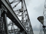 A large steel structure with intricate metal beams and rivets creates an industrial and geometric pattern. The overcast sky provides a backdrop, enhancing the contrast between the shadows and the metallic surfaces. Barbed wire spirals upwards adjacent to the structure, adding an element of tension or security.
