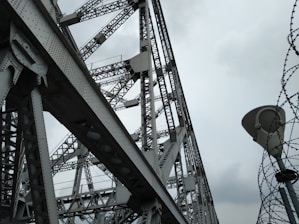 A large steel structure with intricate metal beams and rivets creates an industrial and geometric pattern. The overcast sky provides a backdrop, enhancing the contrast between the shadows and the metallic surfaces. Barbed wire spirals upwards adjacent to the structure, adding an element of tension or security.