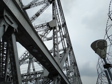 A large steel structure with intricate metal beams and rivets creates an industrial and geometric pattern. The overcast sky provides a backdrop, enhancing the contrast between the shadows and the metallic surfaces. Barbed wire spirals upwards adjacent to the structure, adding an element of tension or security.