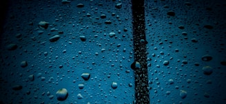 Water droplets cover a transparent surface with a dark vertical line running through the center. The background is a deep blue, accentuating the clear and reflective nature of the droplets.