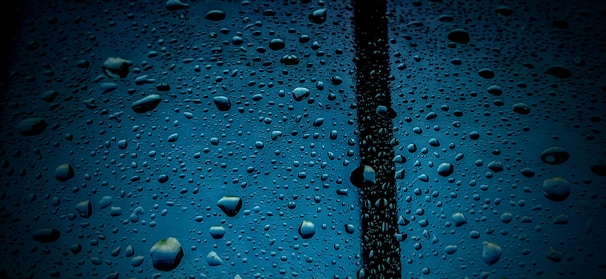 Water droplets cover a transparent surface with a dark vertical line running through the center. The background is a deep blue, accentuating the clear and reflective nature of the droplets.
