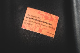 A small rectangular orange paper with a printed quote is attached to a dark surface. The quote is in German and is attributed to Gilles Deleuze. The background of the paper features a subtle pattern resembling floral or organic shapes.