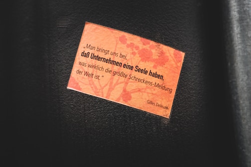 A small rectangular orange paper with a printed quote is attached to a dark surface. The quote is in German and is attributed to Gilles Deleuze. The background of the paper features a subtle pattern resembling floral or organic shapes.