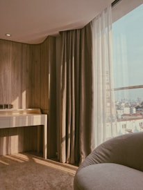 A corner of a room with a soft, warm aesthetic featuring a wooden wall, a light beige vanity table, and large beige curtains. The sunlight streams gently through the window, casting shadows on the carpet. Outside the window, a cityscape with various buildings under a blue sky is partially visible.