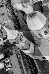 An aerial black and white photograph of an architectural scene featuring a large dome and a smaller tower among densely packed buildings. The rooftop tiles and geometric shapes dominate the image, creating a complex interplay between light and shadow.