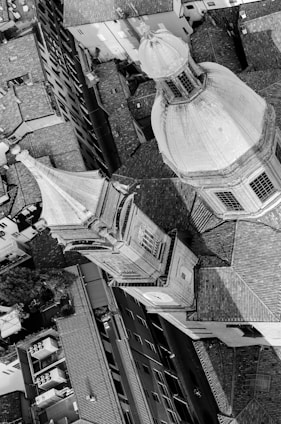 An aerial black and white photograph of an architectural scene featuring a large dome and a smaller tower among densely packed buildings. The rooftop tiles and geometric shapes dominate the image, creating a complex interplay between light and shadow.
