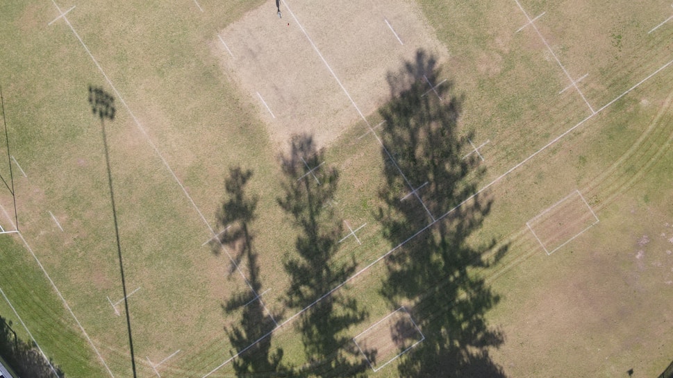 An aerial view of a sports field with marked lines, visible goalposts, and a large shadow cast by nearby trees. The field is mostly grass with patches of dirt and dry areas. The shadow of a floodlight pole is also visible on the left side of the field.