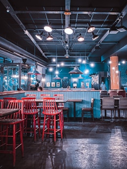 A modern, stylish bar with red high stools around wooden tables. The space features a unique lighting setup with industrial-style pendant lights hanging from the ceiling, creating a warm and inviting atmosphere. The walls are painted a vibrant blue, and there's contemporary art displayed throughout.