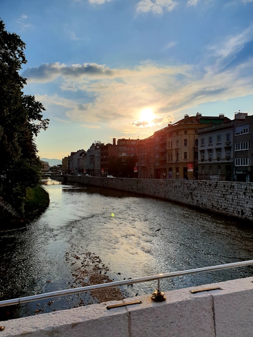 The image features a tranquil river flowing through a city with buildings lining its banks. The sun is setting in the background, casting a golden glow over the scene. The sky is partly cloudy with patches of blue visible. Trees are present on the left side, and there is a bridge visible in the foreground.