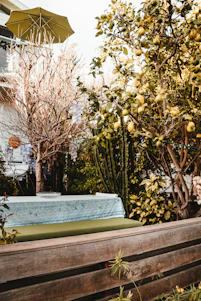 A sunlit patio featuring a wooden bench stool, a woven decorative basket, and a sleek outdoor loveseat surrounded by greenery.